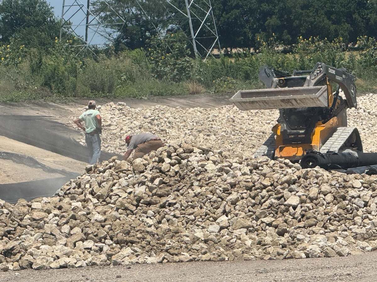Crew hand-setting limestone rip rap over black filter fabric as a skid steer unloads more rock